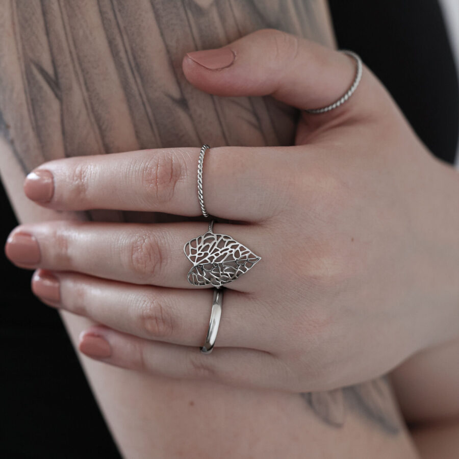 Hopeinen lehtisormus kirurginteräksestä tatuoidulla kädellä vaaleaa taustaa vasten
