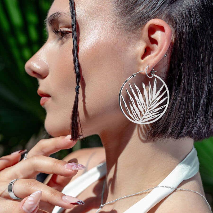 Hopeiset Noble Leaf Hoops korvakorut naisella, jolla on letitetty tukka ja trooppinen tausta