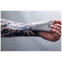 Harmaa-valkoinen No Cosmetics Intensiv Repair Cream -tuubi, jota pitää tatuoitu käsi