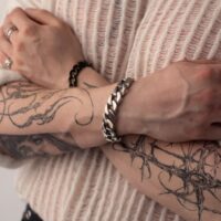 Henkilö, jolla on tatuoidut käsivarret, käyttää hopeista Chunky-rannekorua, kädet ristissä vaalean neuleen päällä