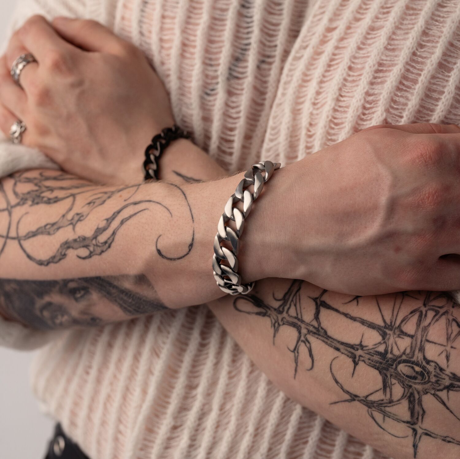 Henkilö, jolla on tatuoidut käsivarret, käyttää hopeista Chunky-rannekorua, kädet ristissä vaalean neuleen päällä