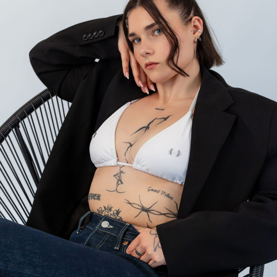 Nainen valkoisessa bikiniyläosassa ja mustassa bleiserissä istuu tuolilla, tatuoitu ylävartalo