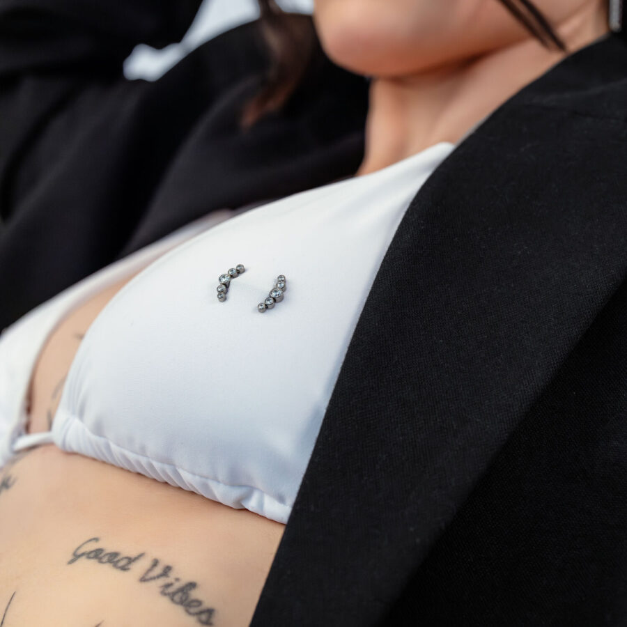 Lähikuva valkoisesta bikinien yläosasta Penta Crystal Barbell -lävistyksellä ja tatuoinnit taustalla.