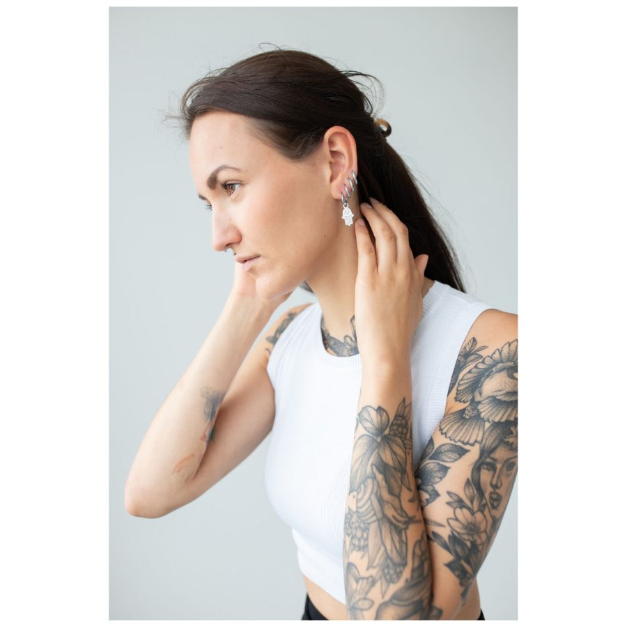 Tatuoitu nainen käyttää Hamsa-korvakoruja, valkoinen toppi, harmaa tausta