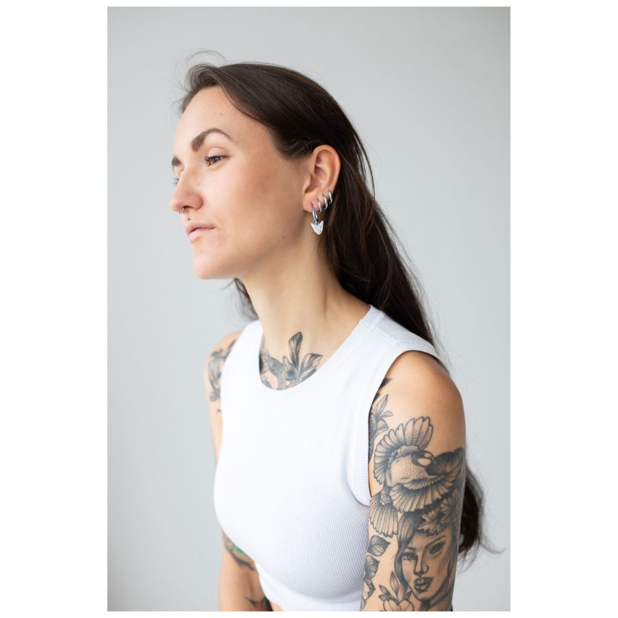 Nainen, jolla on tatuoidut käsivarret, käyttää mustia Little Sphynx Hoopseja harmaata taustaa vasten