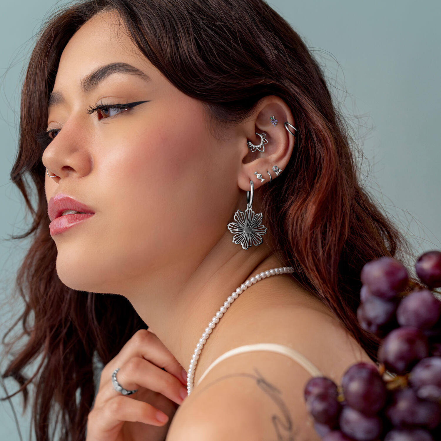 Hopeinen Push Fit Grape -lisäosa Nainen, jolla on useita hopeisia lävistyksiä korvassa, käyttää helmikaulakorua ja pitää rypäleitä.